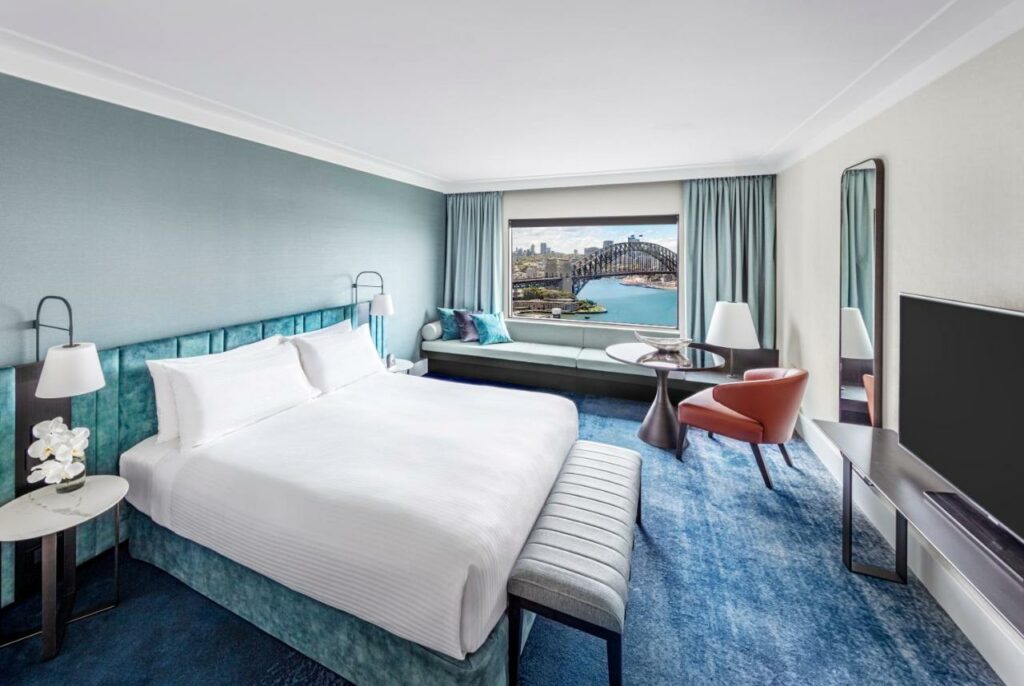 Quarto do InterContinental Sydney, an IHG Hotel. Do lado esquerdo uma cama de casal, com uma mesinha e um abaur de cada ladom na frente cama um sofá. Do lado direito uma TV. No fundo uma mesa com uma poltrona, espelho um sofá e a vista da ponte.