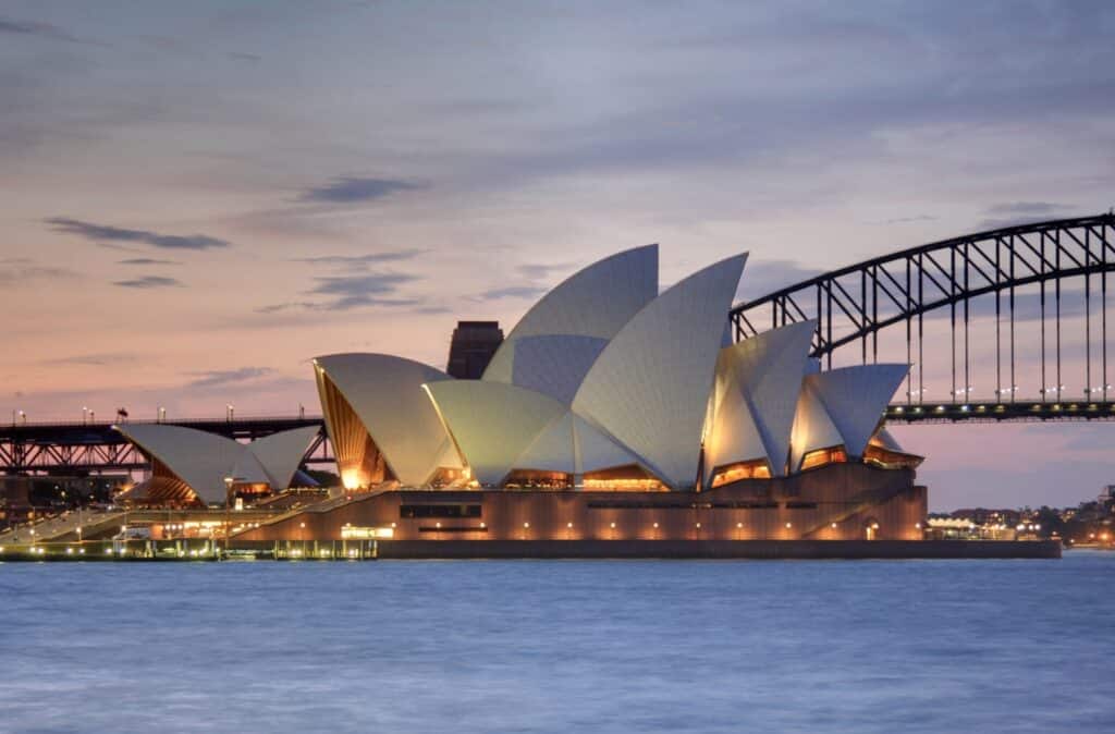 Vista do mar para a Opera House. Uma construção com o teto em formato de conchas e no fundo uma ponte.