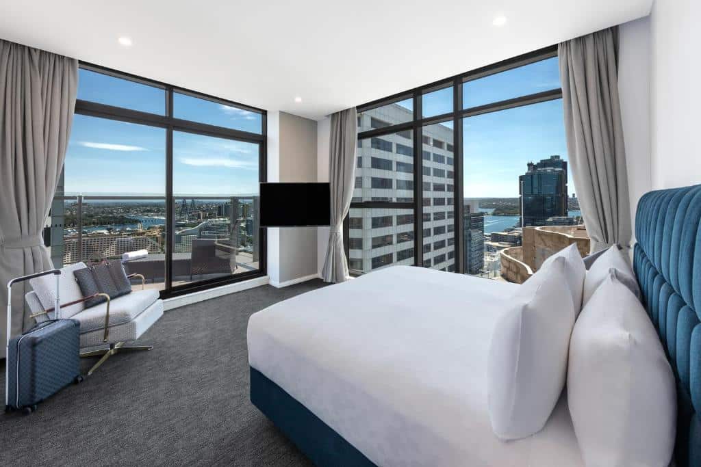 Quarto do Meriton Suites Sussex Street. Do lado direito uma cama de casal, de frente uma poltrona, uma mala e uma TV. No canto esquerdo e no fundo paredes de vidro. Imagem para ilustrar post sobre hotéis em Sydney.