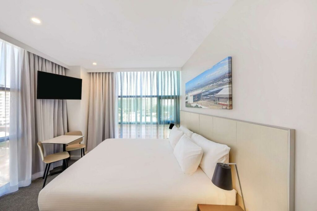 Quarto do Travelodge Hotel Sydney Airport. Do lado direito uma cama de casal, de cada lado uma cômoda com um abajur. Do lado esquerdo uma mesa com duas cadeiras e uma TV. Do lado esquerdo e no fundo paredes de vidro. Imagem para ilustrar post sobre hotéis em Sydney.