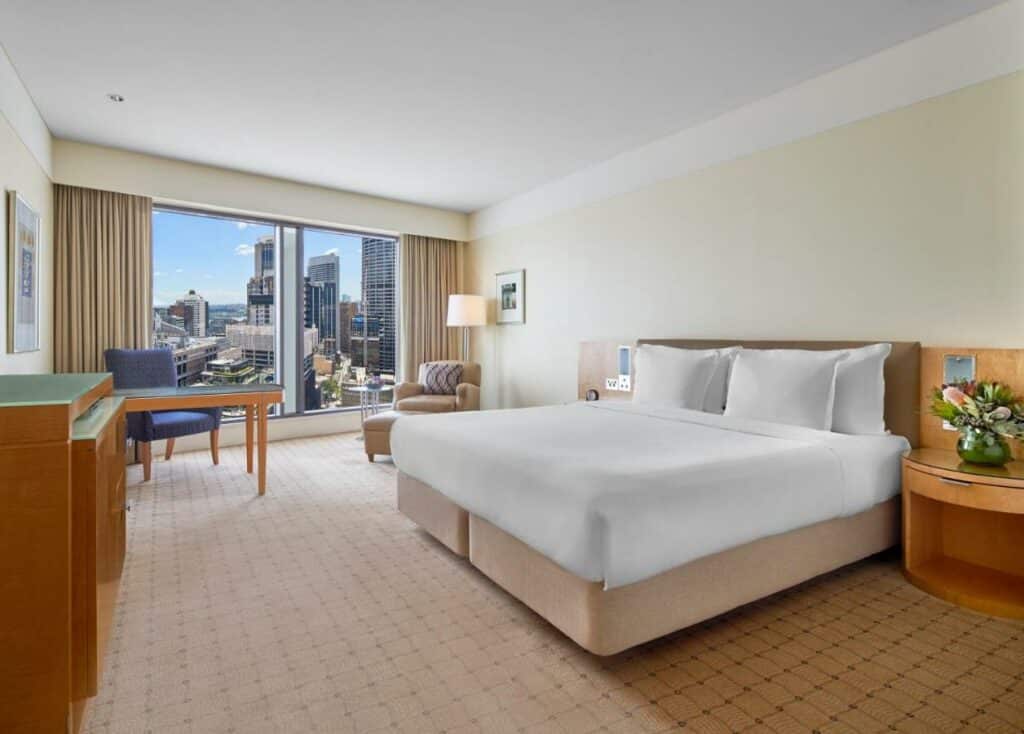 Quarto do The Fullerton Hotel. Do lado direito uma cama de casal, de cada lado uma cômoda, no fundo uma poltrona e um abajur. Do lado esquerdo uma cômoda, no fundo uma mesa de trabalho e uma parede de vidro. Imagem para ilustrar post sobre hotéis em Sydney.