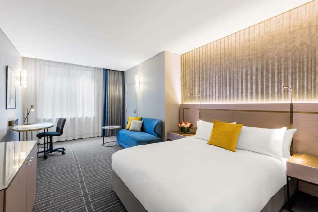 Quarto do Radisson Blu Plaza Hotel. Do lado direito uma cama de casal e uma prateleira com luminária de cada lado. No fundo um pequeno sofá. Do lado esquerdo uma cômoda e uma mesa de trabalho. Imagem para ilustrar post sobre hotéis em Sydney.