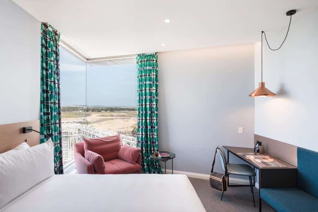 Quarto do Citadines Connect Sydney Airport. Uma cama de casal, uma poltrona e uma parede de vidro do lado esquerdo. Do lado direito uma mesa de trabalho e um sofá. Imagem para ilustrar post sobre hotéis em Sydney.