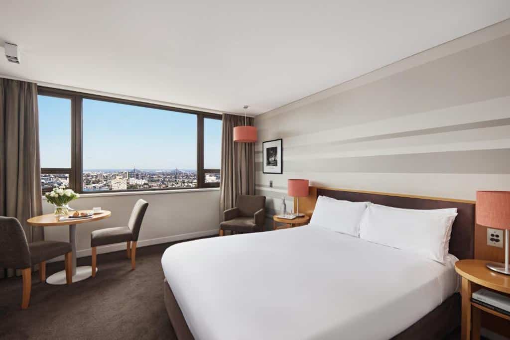 Quarto do View Sydney. Do lado direito uma cama de casal, de cada lado uma cômoda com um abajur, no fundo uma poltrona. Do lado esquerdo uma mesa com duas cadeiras, um vaso e atrás a janela. Imagem para ilustrar post sobre hotéis em Sydney.