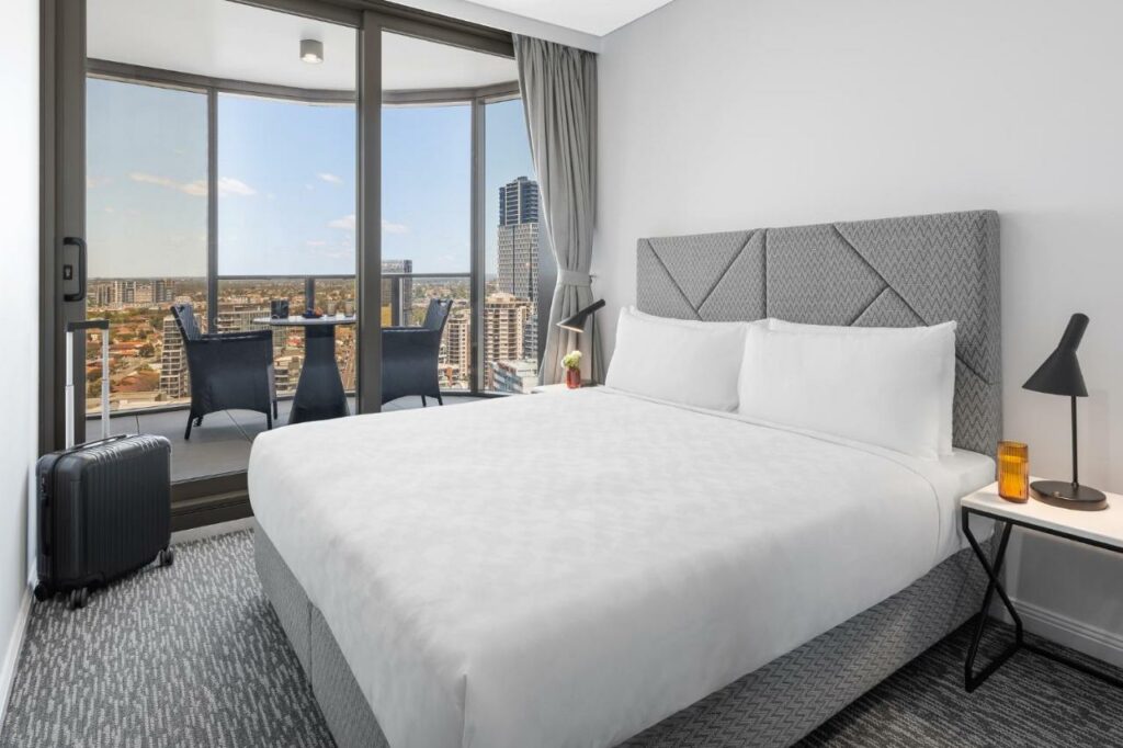 Quarto do Meriton Suites George Street, Parramattael. Uma cama de casal do lado direito, de cada lado uma mesinha com um abajur. Do lado esquerdo uma mala. No fundo a varanda do quarto com uma mesa e duas cadeiras. Imagem para ilustrar post sobre hotéis em Sydney.