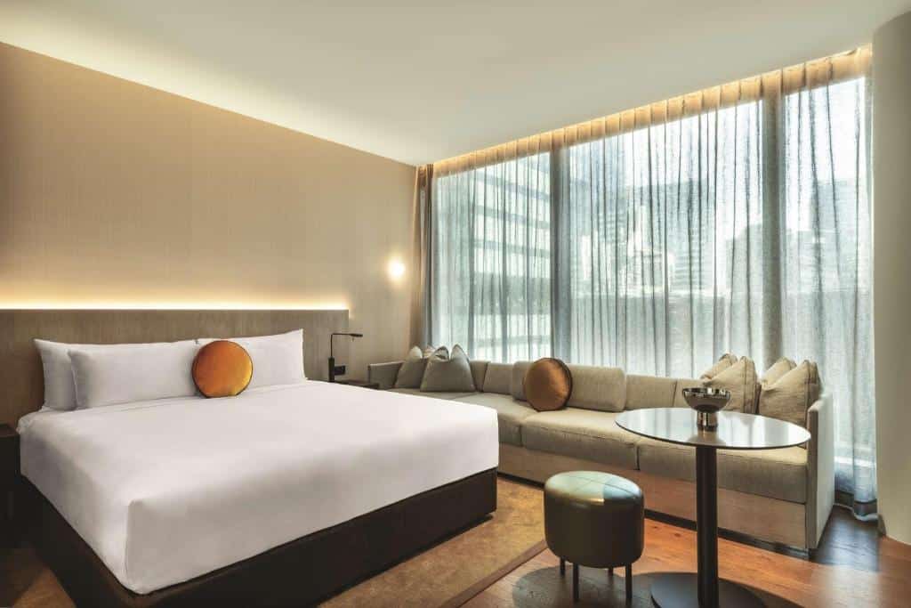 Quarto do A by Adina. Do lado esquerdo uma cama de casal, do lado direito uma mesa com um puff. No fundo um sofá e atrás uma parede de vidro. Imagem para ilustrar post sobre hotéis em Sydney.