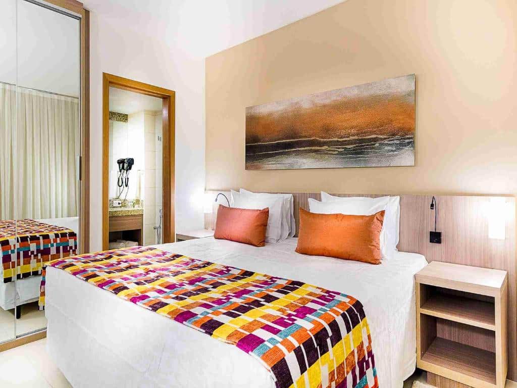 Quarto do Quality Hotel Pampulha & Convention Center com cama de casal, com duas cômodas ao lado com luminárias, porta do banheiro do lado esquerdo e espelho grande ao lado da porta.