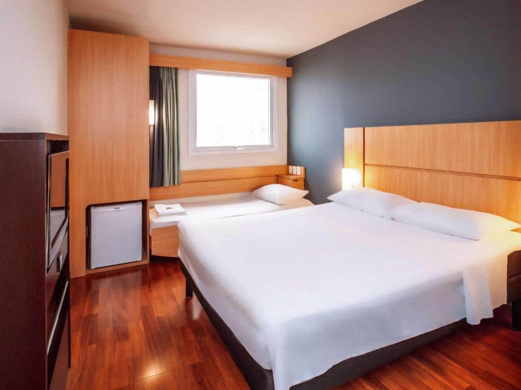 Quarto do Ibis Belo Horizonte Savassi com cama de casal e do lado esquerdo abaixo da janela há uma cama de solteiro com frigobar ao lado e cômoda com TV em frente a cama de casal.