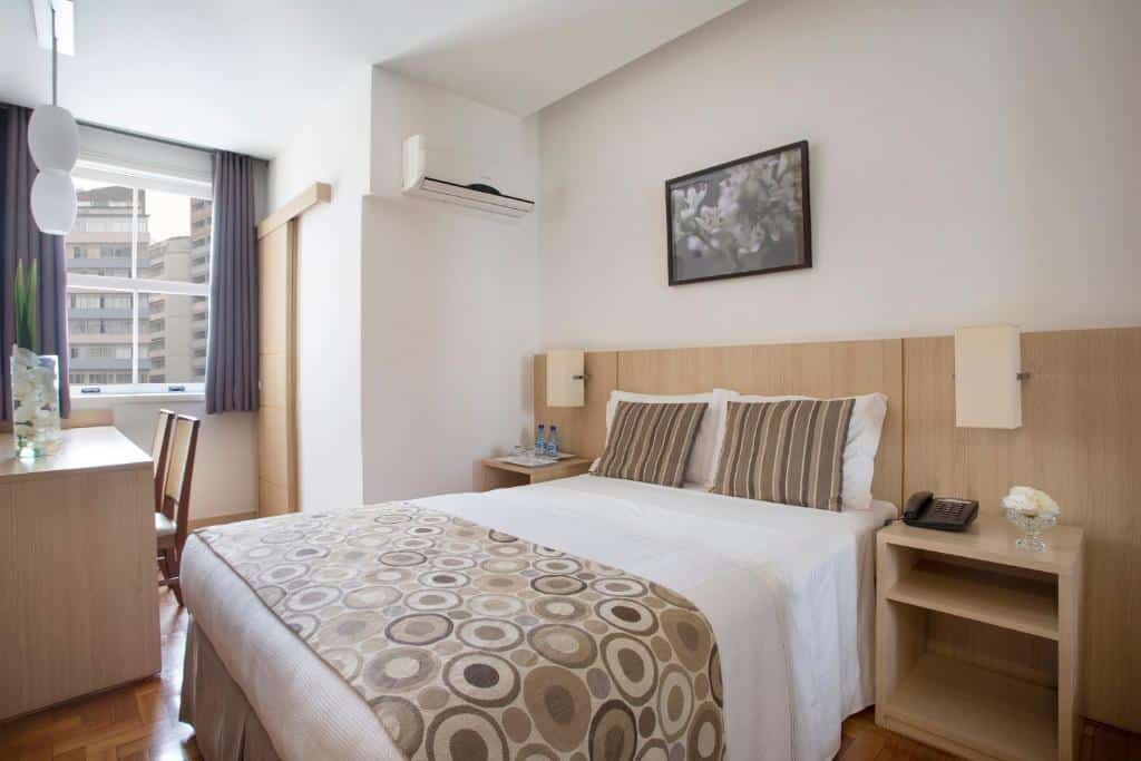 Quarto do Normandy Hotel  com cama de casal, duas cômodas ao lado, sendo a cômoda do lado esquerdo com telefone. Mesa de trabalho com cadeira do lado direito perto da janela com cortinas azuis.