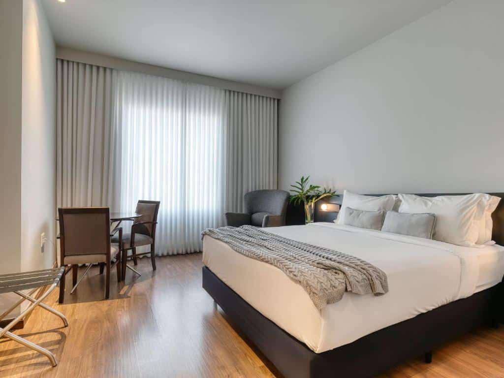 Quarto do Mercure Belo Horizonte Lourdes  com cama de casal, do lado esquerdo um vaso de planta em cima de uma cômoda, poltrona cinza ao lado e a frente da poltrona uma mesa de madeira redonda com duas cadeiras de madeiras com estofado bege.