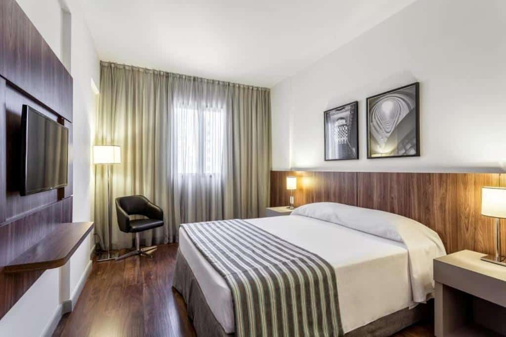 Quarto do Hotel Vivenzo Savassi Belo Horizonte com cama de casal, duas cômodas ao lado da cama, janela do lado esquerdo com cortinas bege, poltrona preta e TV em frente a cama. 