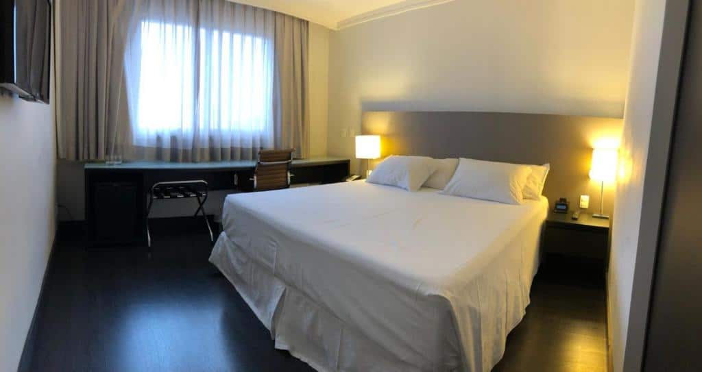 Quarto do Hotel Gran Vivenzo Belo Horizonte com cama de casal, duas luminárias ao lado da cama, mesa de trabalho com cadeira do lado esquerdo em frente a janela com cortinas. Em frente a cama tem uma TV de tela plana.