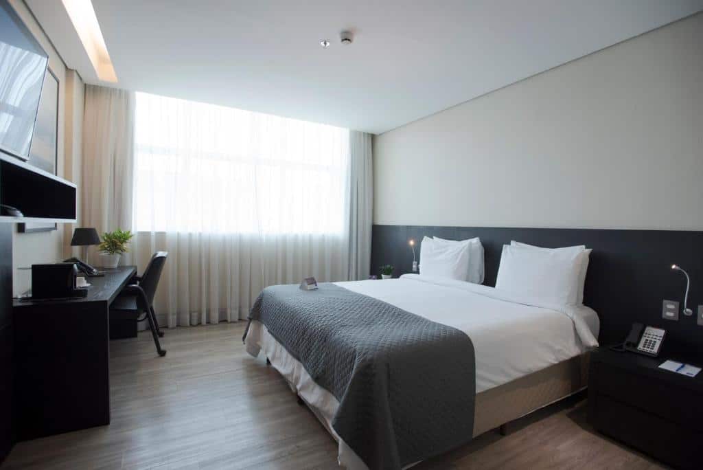 Quarto do Tryp by Wyndham Belo Horizonte Savassi com cama de casal, duas cômodas ao lado da cama, janela do lado esquerdo, mesa de trabalho em frente a cama com TV a cima na parede.