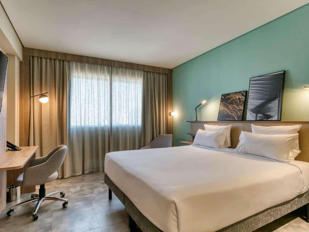 Quarto do Novotel BH Savassi com cama de casal, janela do lado esquerdo com cortinas cinzas, cadeira no lado esquerdo e mesa de trabalho com cadeira em frente a cama.