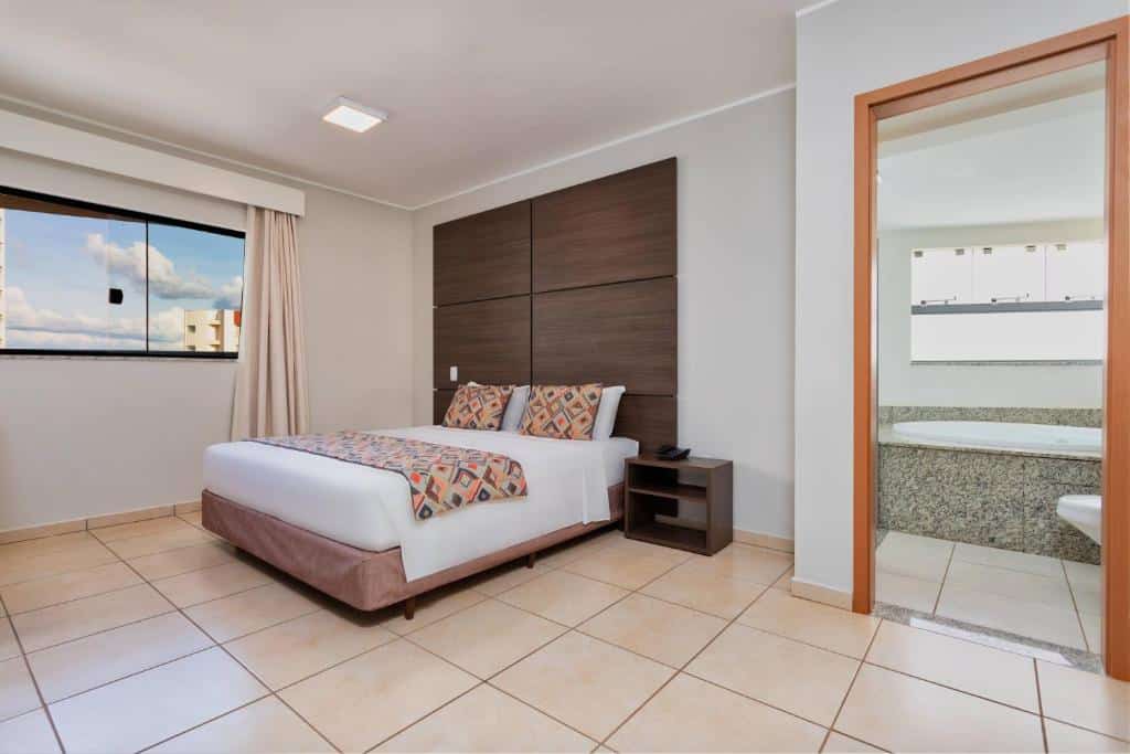 suíte do Wyndham Olímpia Royal Hotels com uma cama de casal, dois travesseiros estampados, uma mesa de cabeceira, uma janela grande e uma porta de banheiro aberta.