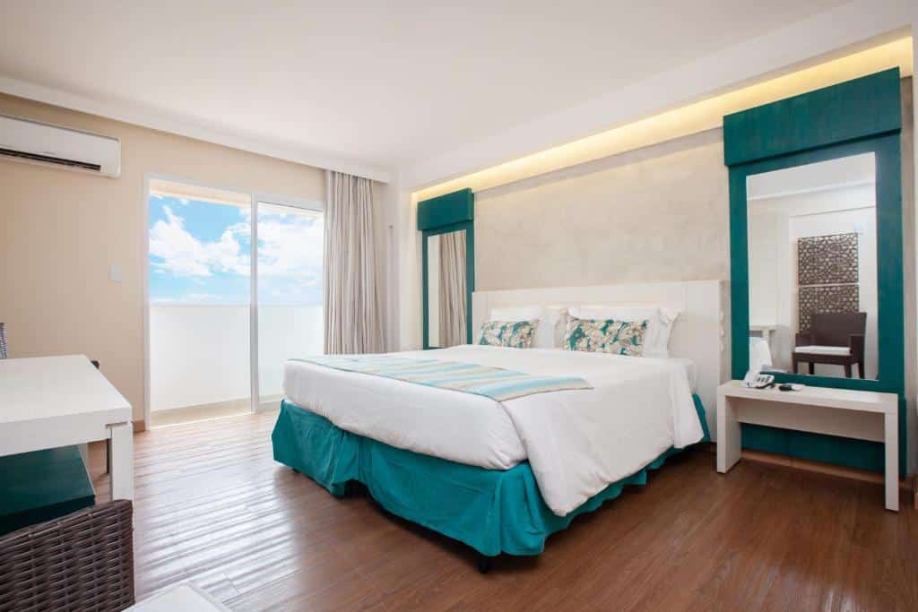 quarto moderno do Celebration Resort Olimpia com cama de casal, detalhes em turquesa, piso de madeira, escrivaninha e varanda com vista para o céu.