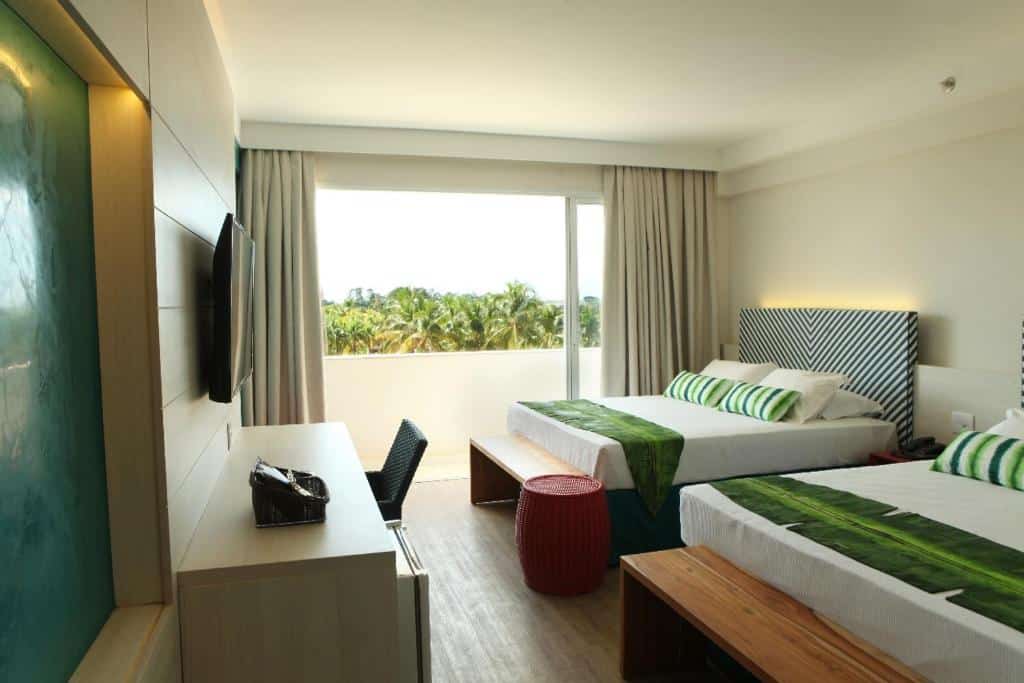 quarto do Hot Beach Resort com duas camas de casal, uma escrivaninha, TV e uma grande janela com vista para as palmeiras do lado de fora.