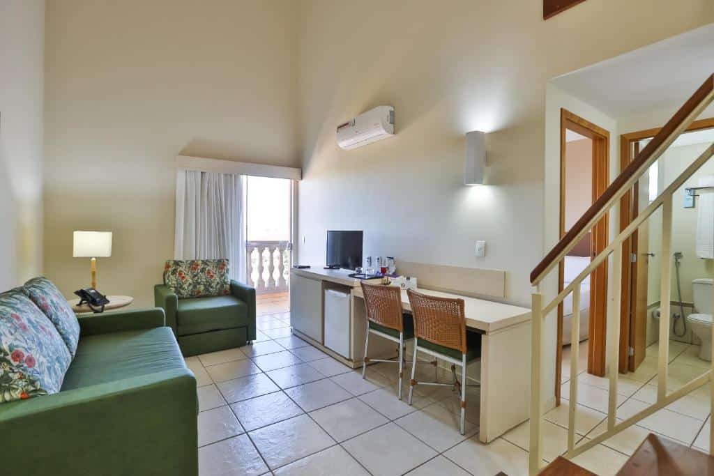 flat do Thermas de Olimpia Resorts by Mercure mostrando um quarto de hotel com piso de azulejos, sofá verde, mesa com cadeiras, TV, escada e vista para o banheiro e a varanda.