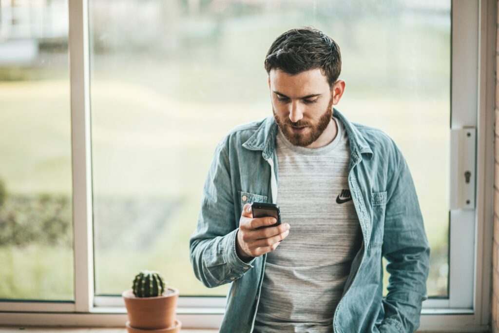 Homem com o cabelo e barba castanho escuro e pele branca, vestido de camiseta jeans aberta, encostado em um balcão com um vaso de cacto ao lado, segurando o celular com uma mão. Ao fundo tem uma janela de vidro com natureza
