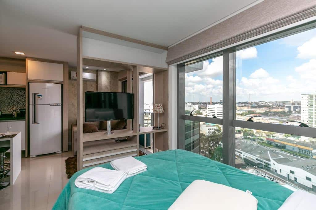 Um moderno apartamento estúdio com cama, TV, área de cozinha e grandes janelas que mostram a paisagem urbana sob um céu azul, no Conforto e Comodidade - Studio Central - 7th019, para representar airbnb em Curitiba.