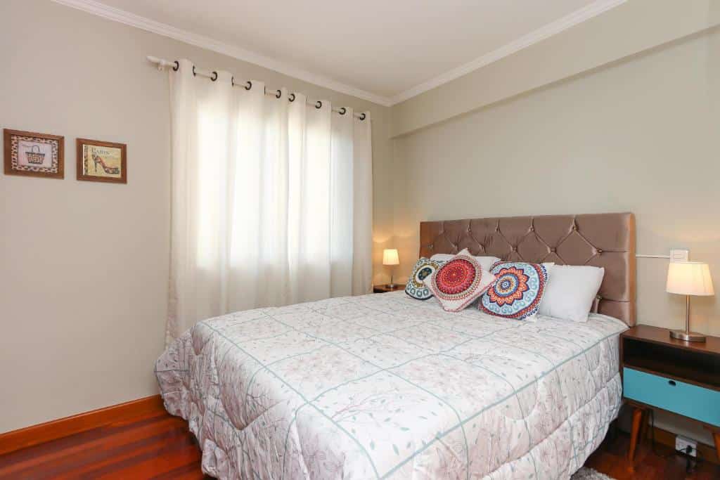 Uma cama bem arrumada com travesseiros estampados, duas mesas de cabeceira com abajures, arte emoldurada e uma janela com cortinas brancas, no Lindo Apartamento - Vista Parque Barigui, para representar airbnb em Curitiba.