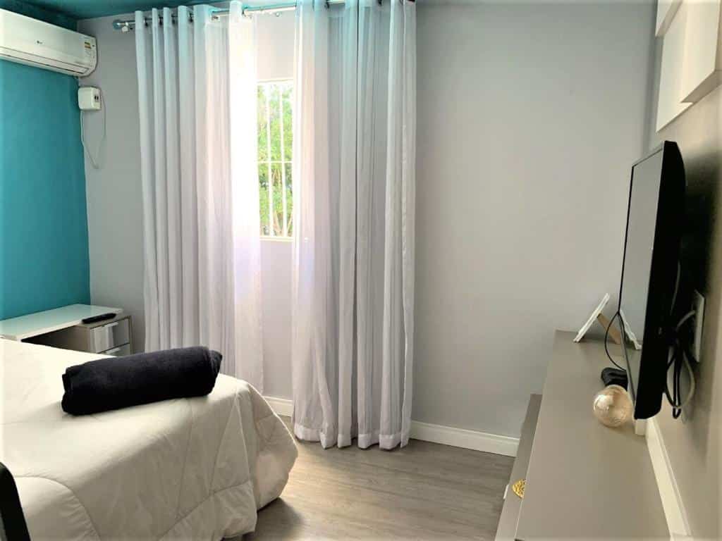 Um quarto com uma cama, uma TV em um console, uma parede azul, cortinas brancas e uma janela que deixa entrar a luz do dia, na Mello's House, para representar airbnb em Curitiba.
