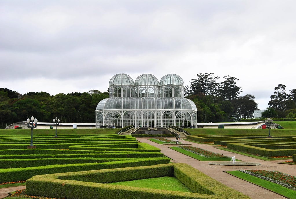 Grande estufa de vidro com cúpulas, cercada por jardins bem cuidados e cercas vivas sob um céu nublado, trata-se do Palácio no Jardim Botânico em Curitiba.