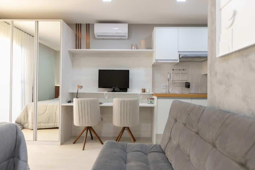 Estúdio moderno com uma área de trabalho, duas cadeiras, uma TV na parede e um sofá cinza em primeiro plano, no Studio A Lenzi - AYN062, para representar airbnb em Curitiba.