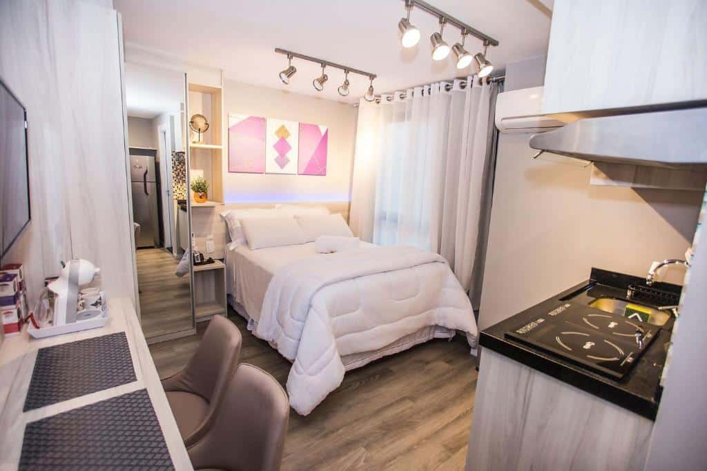 Estúdio moderno com cama, escrivaninha, cozinha pequena, arte na parede e janela grande com cortinas transparentes, do Studio Completo no centro de Curitiba - ALL1021, para representar airbnb em Curitiba.