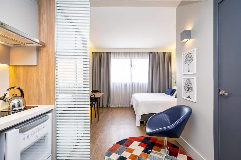 Estúdio moderno com uma cama, uma pequena mesa de jantar, cadeira e cozinha compacta, iluminado por luz natural de uma janela, no Helbor Stay Batel - HSB033, para representar airbnb em Curitiba.