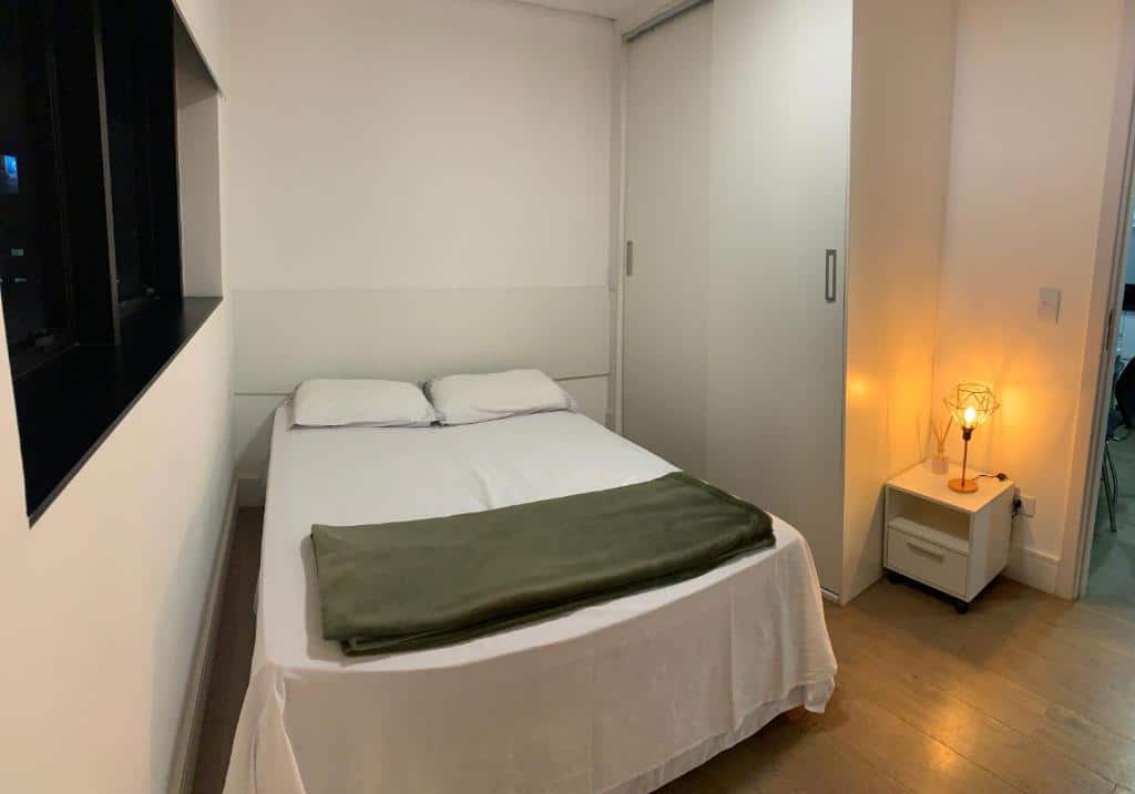 Uma cama bem arrumada com lençóis brancos e um cobertor verde fica ao lado de uma mesa de cabeceira com uma lâmpada acesa em um quarto pequeno, no Lindo Apartamento 709, para representar airbnb em Curitiba.