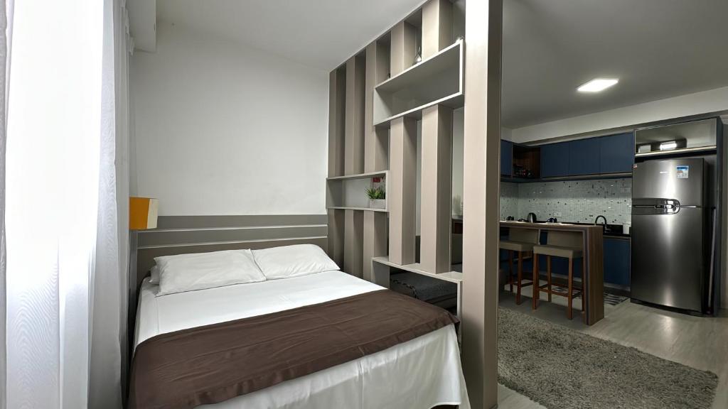 Um pequeno quarto com uma cama de casal, divisória parcial e vista para uma área de cozinha com geladeira e cadeiras de balcão, no Studio Batel, para representar airbnb em Curitiba.