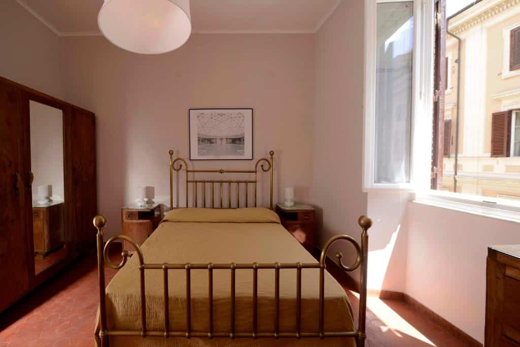 quarto do Via Montebello um airbnb em Roma, com decoração rústica, cama de casal, mesinha de cabeceira com luminária de ambos os lados, com janela de um lado e, do outro lado, guarda-roupa com espelho em madeira, e luz também no teto