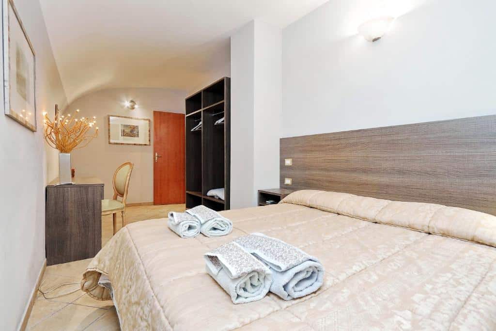 quarto do Monti House Rome, um airbnb em Roma, cama bem grande, toalhas em cima, mesinha de cabeceira de ambos os lados, guarda-roupa aberto e, em frente, mesa com cadeira 