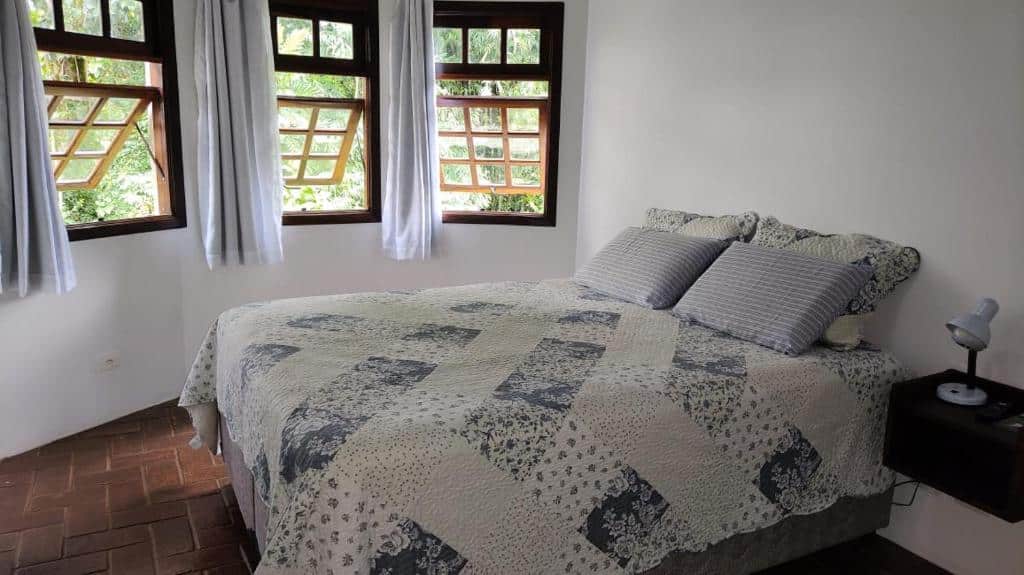 Foto do quarto do Centro – São Francisco Xavier – Casa Agradável. Representa o post sobre airbnb em São Francisco Xavier. A cama é de casal e ao lado direito dela há uma mesa de cabeceira com abajur, enquanto à esquerda há três janelas.
