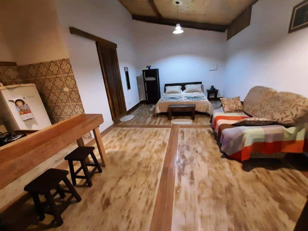 Um quarto aconchegante do chalé Filhos da Terra com uma cama, sofá, piso de madeira, pequena área de cozinha, parede estampada e bancos ao lado de um balcão de madeira.