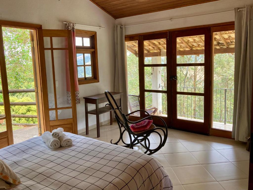 Quarto do Chalés de Valfena com colcha xadrez, cadeira de balanço de madeira e grandes janelas com vista para uma área externa verdejante.