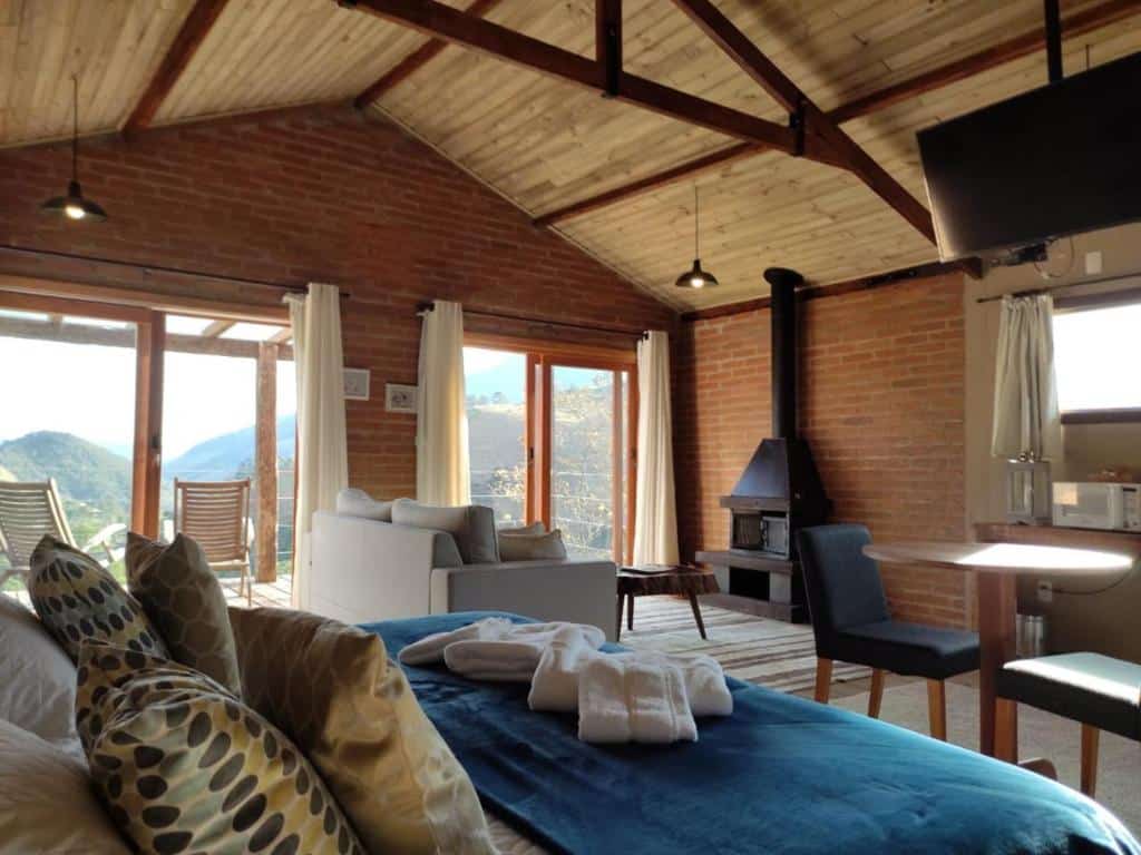 Foto do quarto da Pousada Serra do Luar. Representa o post sobre airbnb em São Francisco Xavier. A Cama está no canto esquerdo. Na frente dela há uma mesa com duas cadeiras, e também vemos parte da cozinha. Ao fundo da foto há uma sala com sofá e lareira, e mais ao fundo há portas e janelas de vidro com vista.