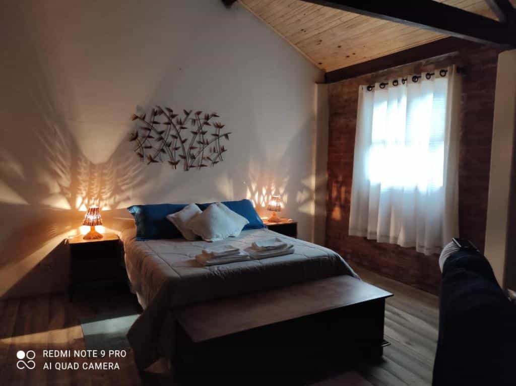 Quarto aconchegante do Chalé Lavanda com Hidro com uma cama de casal, duas mesas de cabeceira com luminárias, uma janela com cortinas transparentes e um móvel decorativo de parede acima da cama.