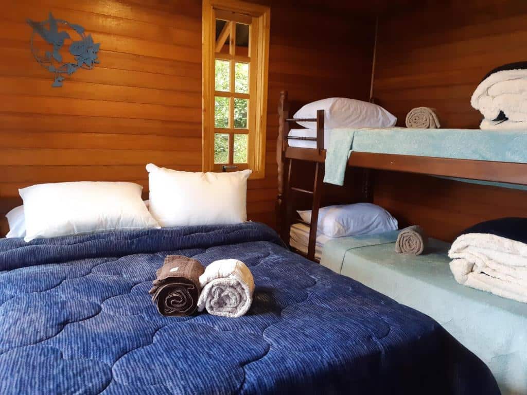 A imagem mostra o interior de um quarto pequeno e aconchegante, com paredes de madeira em tom claro no Chalé Beija-Flor. Há uma cama de casal com colcha azul escura e travesseiros brancos, com toalhas enroladas em cima da cama. Ao lado da cama, há um beliche de madeira com dois colchões, cada um coberto por uma colcha azul clara. O beliche tem escada simples de madeira e mais toalhas dobradas estão dispostas na cama superior. Ao fundo, há uma janela de madeira que permite a entrada de luz natural, iluminando o ambiente.