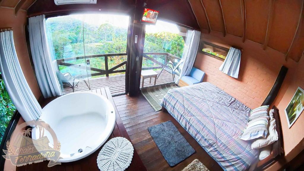 A imagem mostra o interior de um dos Chalés do Ferradura aconchegante com um quarto espaçoso. À esquerda, há uma banheira de hidromassagem branca junto a uma grande janela de vidro que oferece vista para a natureza externa. Ao lado da janela, uma porta de vidro deslizante leva a uma pequena varanda com duas cadeiras e uma mesa, voltada para a paisagem verde. No lado direito do quarto, há uma cama de casal com roupa de cama em tons neutros e travesseiros organizados. O chão é de madeira, e o ambiente tem uma atmosfera acolhedora, iluminado pela luz natural que entra pelas janelas.