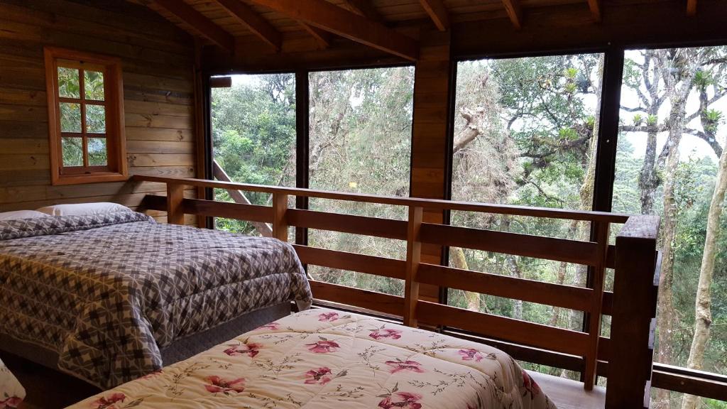 A imagem mostra o interior de um quarto do Chalé dos tucanos com paredes e teto de madeira, oferecendo uma vista ampla da floresta através de grandes janelas de vidro. O quarto contém duas camas, uma maior com colcha cinza e branca em padrão geométrico e outra menor com colcha floral em tons de rosa. Ao lado das camas, há uma sacada interna com grades de madeira que dá vista para a natureza ao redor. A janela lateral de madeira permite a entrada de luz natural, criando um ambiente acolhedor e imerso na vegetação ao redor.