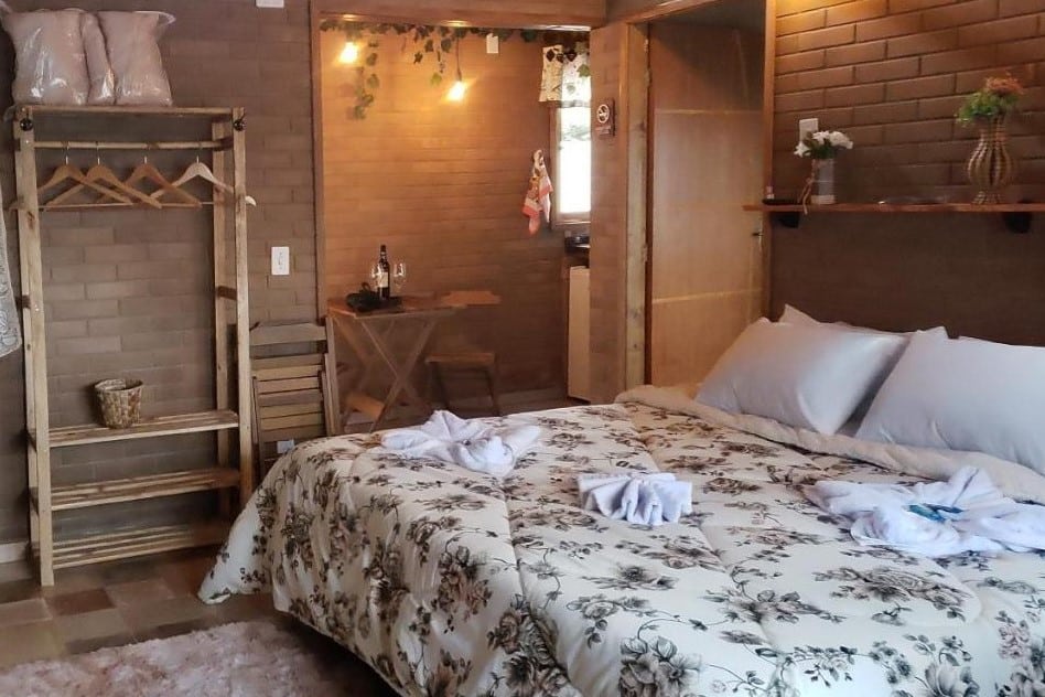 A imagem mostra um quarto aconchegante com paredes de tijolos à vista em tons terrosos na Pousada Grom's Village. No centro, há uma cama de casal grande com uma colcha de estampa floral em tons neutros, além de travesseiros brancos. Ao lado da cama, há uma estante de madeira rústica usada como armário, com cabides expostos. No fundo, pode-se ver uma pequena mesa de madeira com uma garrafa de vinho e duas taças, sob uma luz suave, dando um toque romântico ao ambiente. O quarto tem uma decoração simples e acolhedora, com detalhes rústicos.