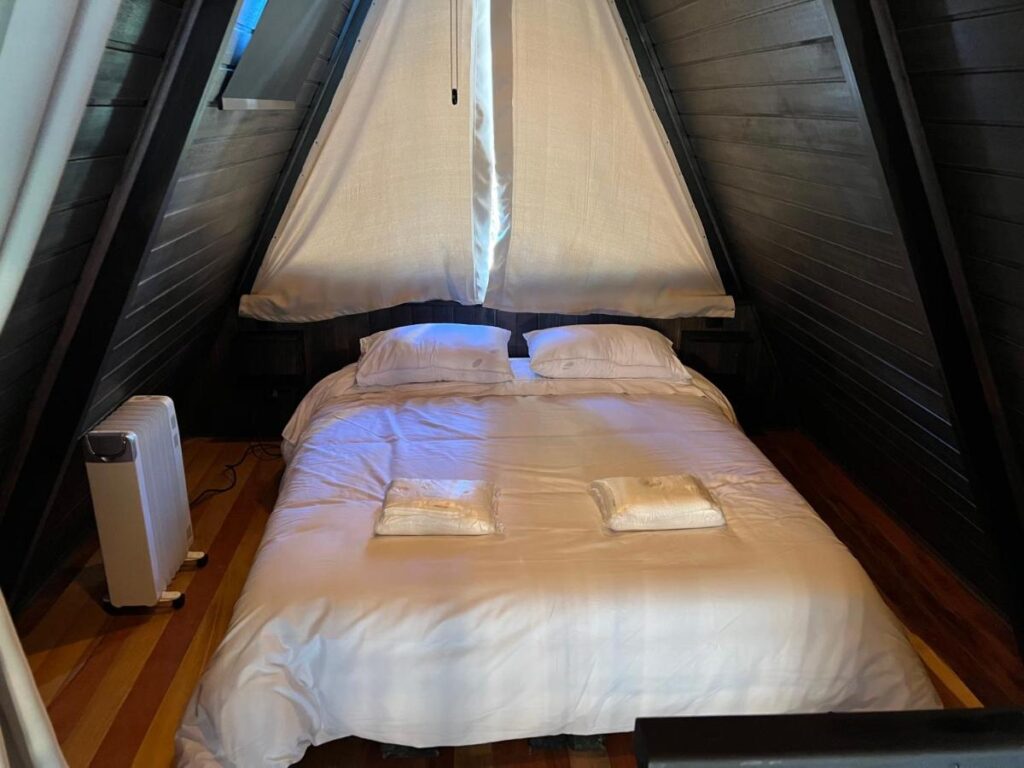 A imagem mostra o interior de um chalé em formato de "A", com um ambiente acolhedor e íntimo no Chalé Villa Nature. No centro, há uma cama de casal com roupa de cama branca, situada sob um teto triangular com paredes de madeira escura. Acima da cama, há uma grande janela triangular coberta por uma cortina branca, que permite a entrada de luz natural. Ao lado da cama, há um pequeno aquecedor para manter o ambiente aquecido. O piso é de madeira clara, contrastando com as paredes escuras, criando uma sensação de conforto e tranquilidade.