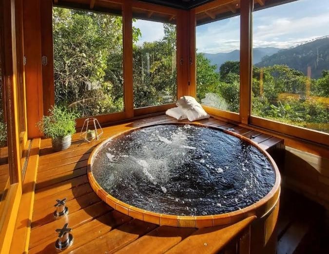 Uma banheira de hidromassagem redonda de madeira com água borbulhante fica perto de grandes janelas, oferecendo uma vista de colinas verdejantes e um céu ensolarado.