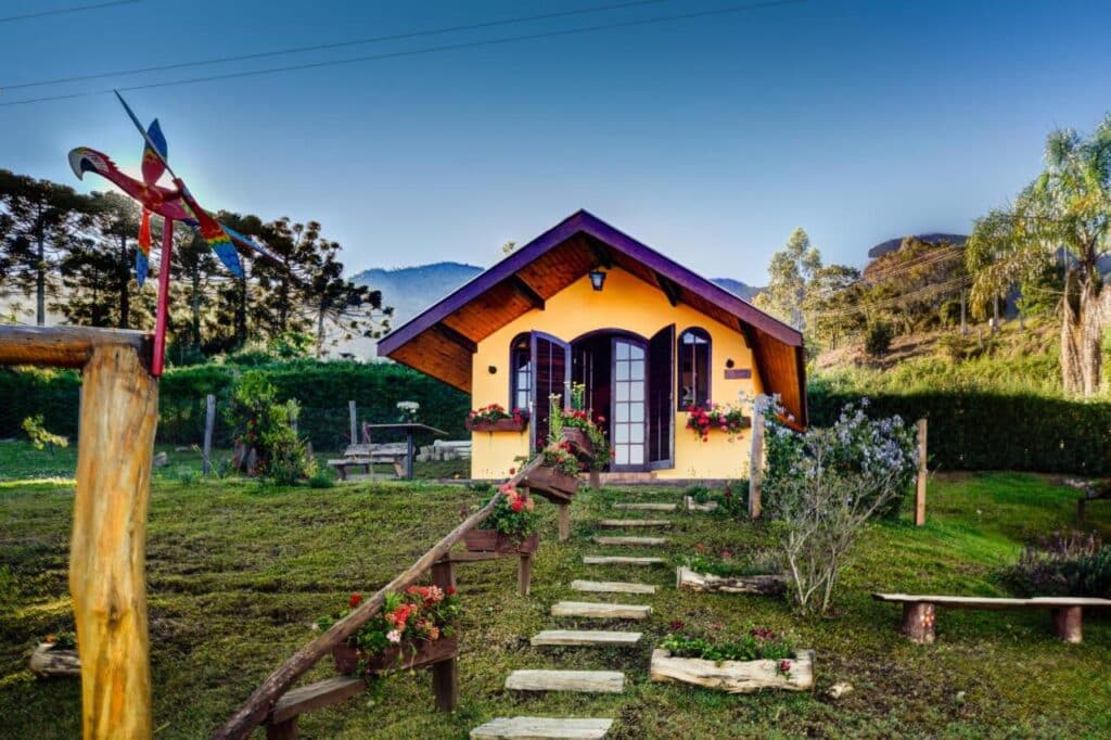 Lado exterior de um dos Chalés Pedra do Baú. É uma casa pequena e colorida com um caminho de madeira, cercada por grama, flores e árvores, com montanhas ao fundo.