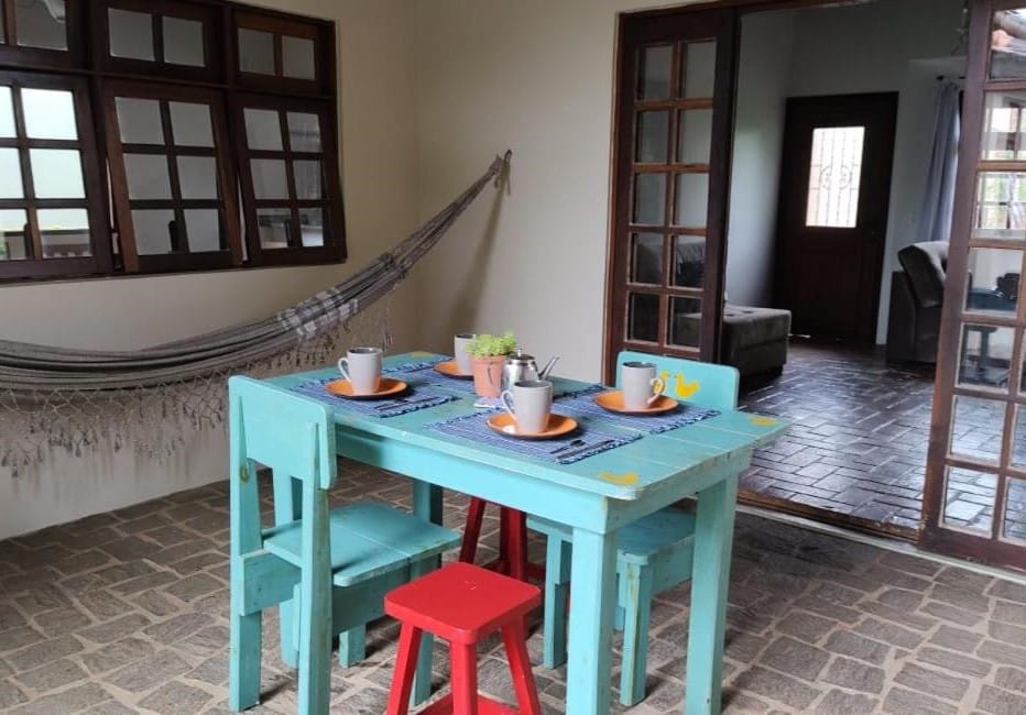 Uma aconchegante área de jantar com uma mesa turquesa, quatro cadeiras descombinadas, jogos americanos e uma rede ao lado de grandes janelas.