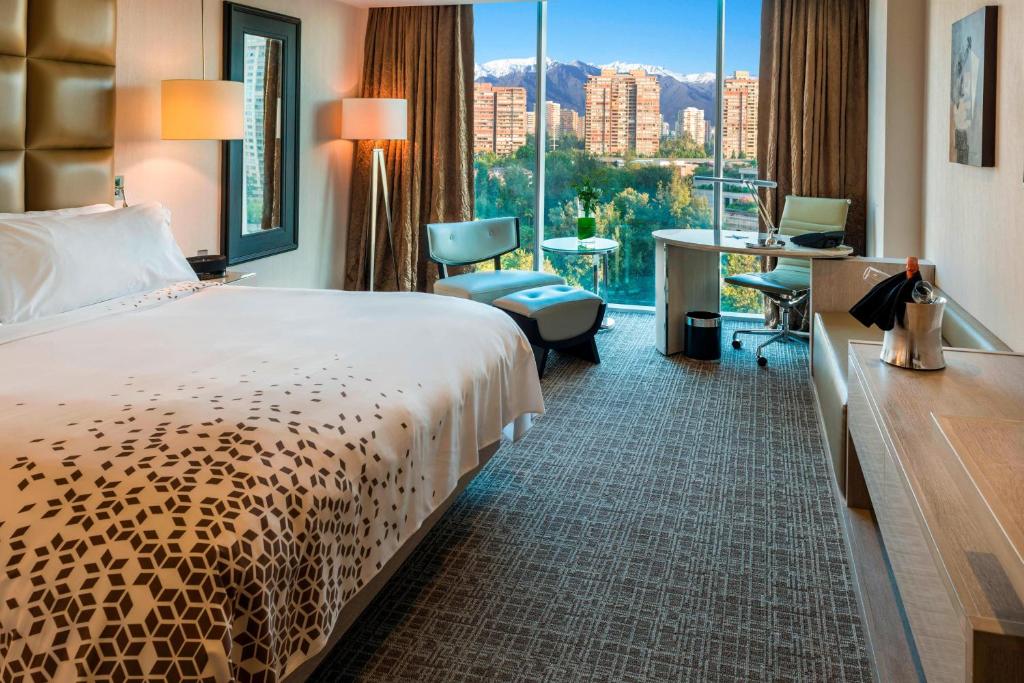 Quarto do Renaissance Santiago Hotel com cama de casal do lado esquerdo e em frente a cama cômoda de madeira e do lado esquerdo do quarto poltrona e uma mesa de trabalho. 