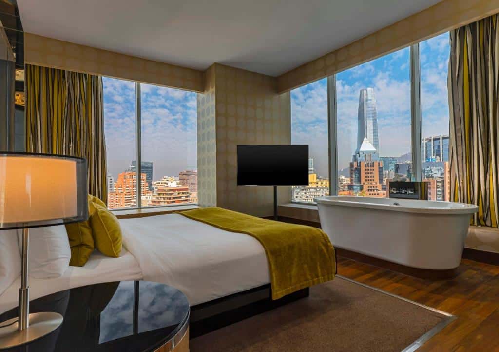 Quarto do W Santiago com cama de casal do lado esquerdo e em frente a cama banheira de hidromassagem ao lado de janelas amplas com vista para a cidade. Representa hotéis 5 estrelas em Santiago.
