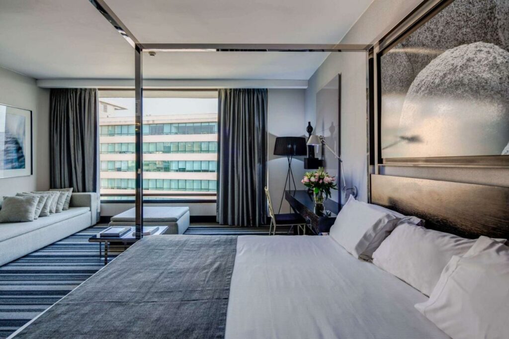 Quarto do Wyndham Santiago Pettra com cama de casal do lado direito da imagem a frente do lado direito da cama ao fundo uma mesa com cadeira e do lado esquerdo um sofá. Representa hotéis 5 estrelas em Santiago.